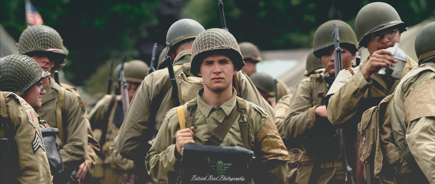 A soldier standing in ranks, staring straight ahead with a focused expression. Dressed in uniform, his posture is straight and disciplined, embodying the ideals of duty and commitment as part of a larger military formation.